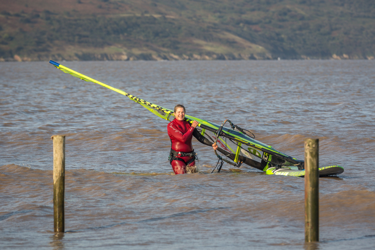 Lady in a red wetsuit standing in the sea holding her windsurfing board and sail